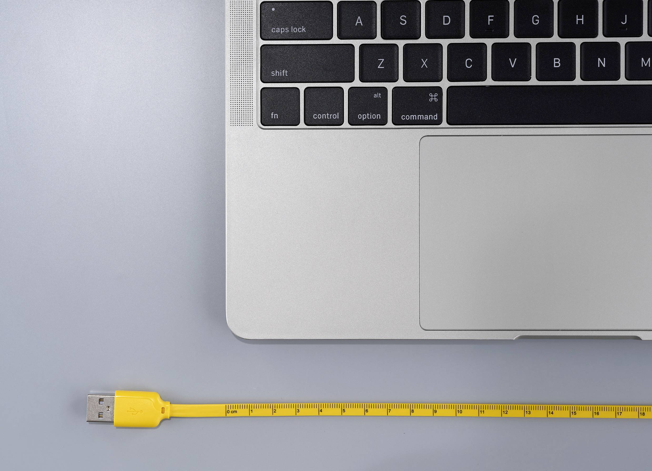 Laptop-toetsenbord in de rechterbovenhoek, geel USB-kabel met centimetermarkeringen eronder op een grijze achtergrond.