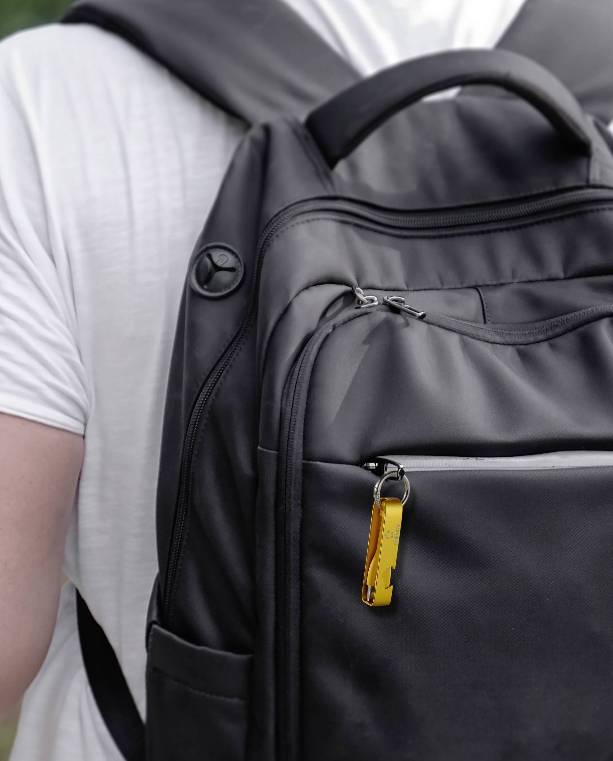 Een persoon draagt een zwarte rugzak. Aan de rits hangt een fel gele sleutelhanger.