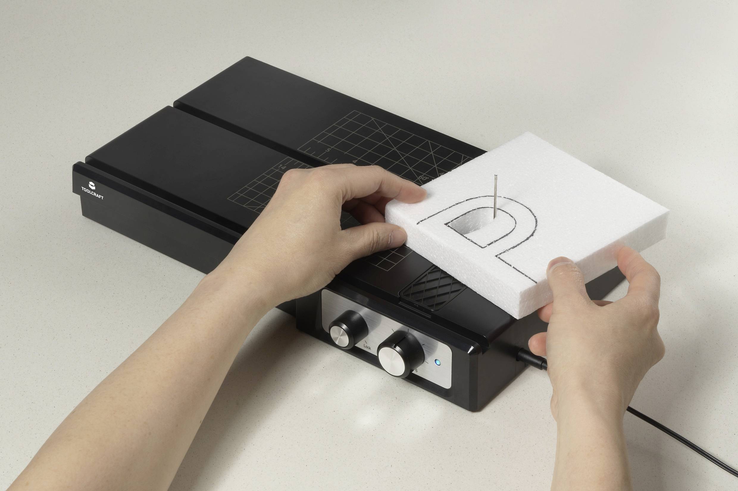 Twee handen snijden met een warmtedraadsnijder een letter 'D' uit wit schuimmateriaal.