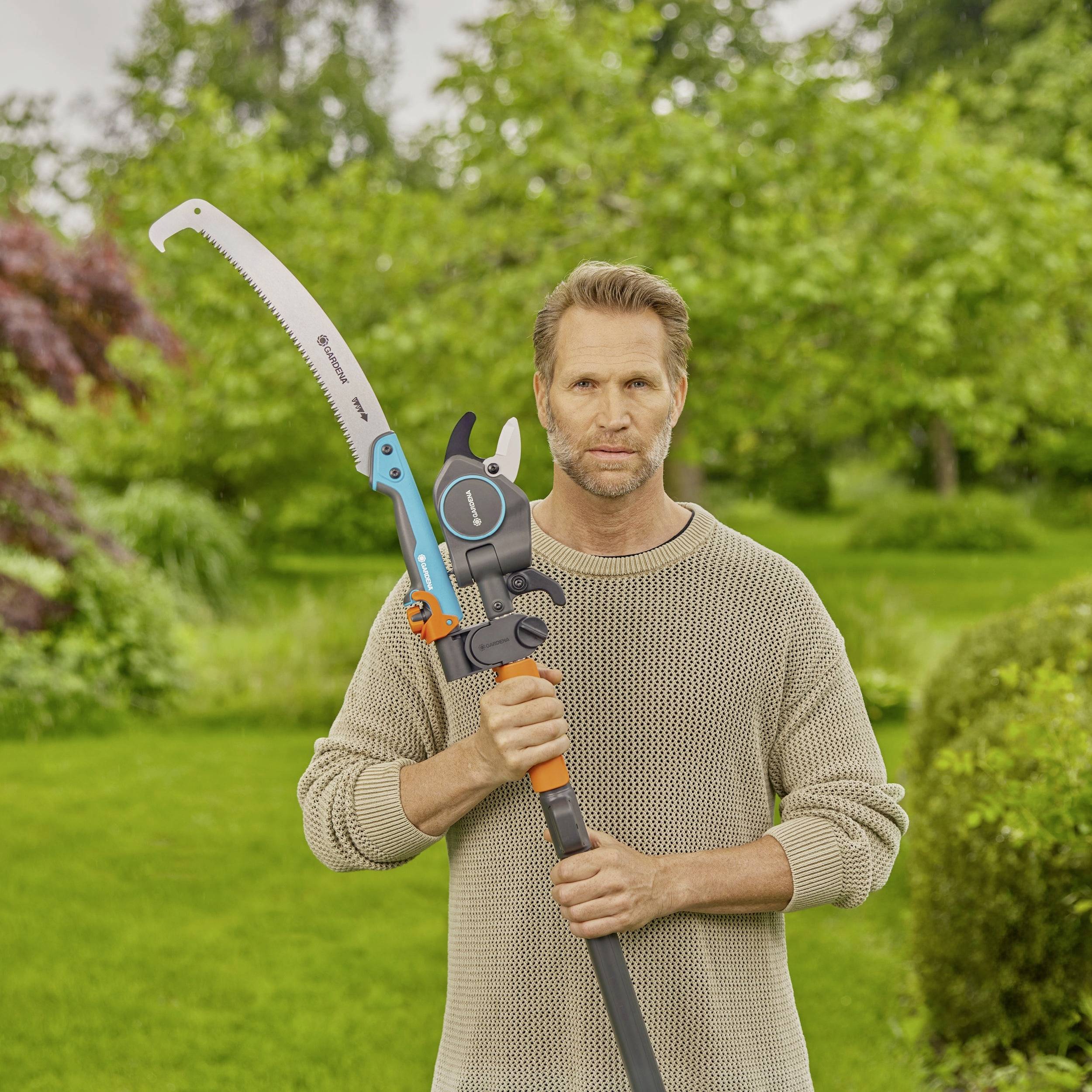 Een man staat in een tuin en houdt een handzaag met oranje en grijze kleuren. Op de achtergrond zijn bomen en struiken zichtbaar.