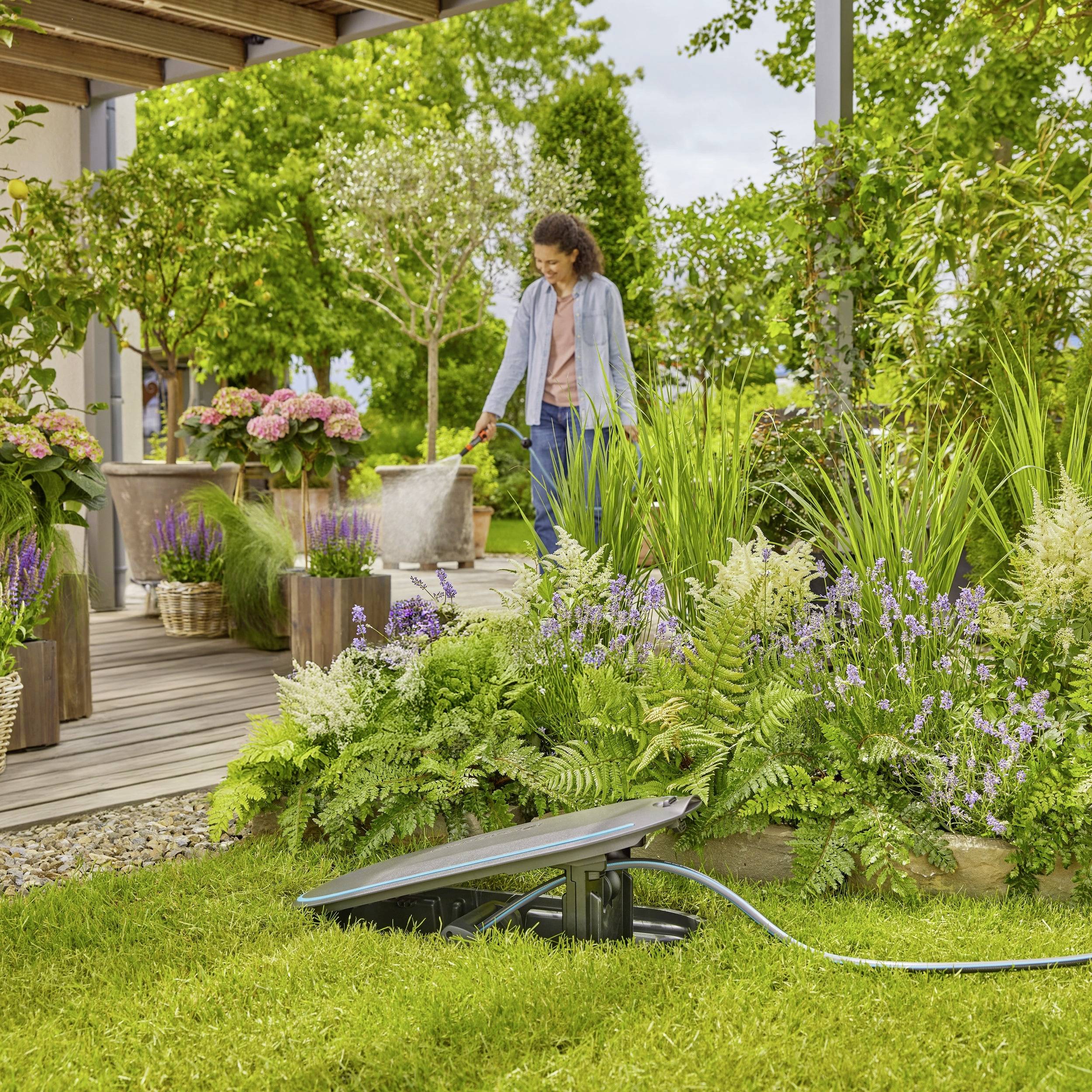 Een persoon staat in een tuin naast bloemen en planten, omringd door groen loof. Een zonnelamp is op de voorgrond zichtbaar.