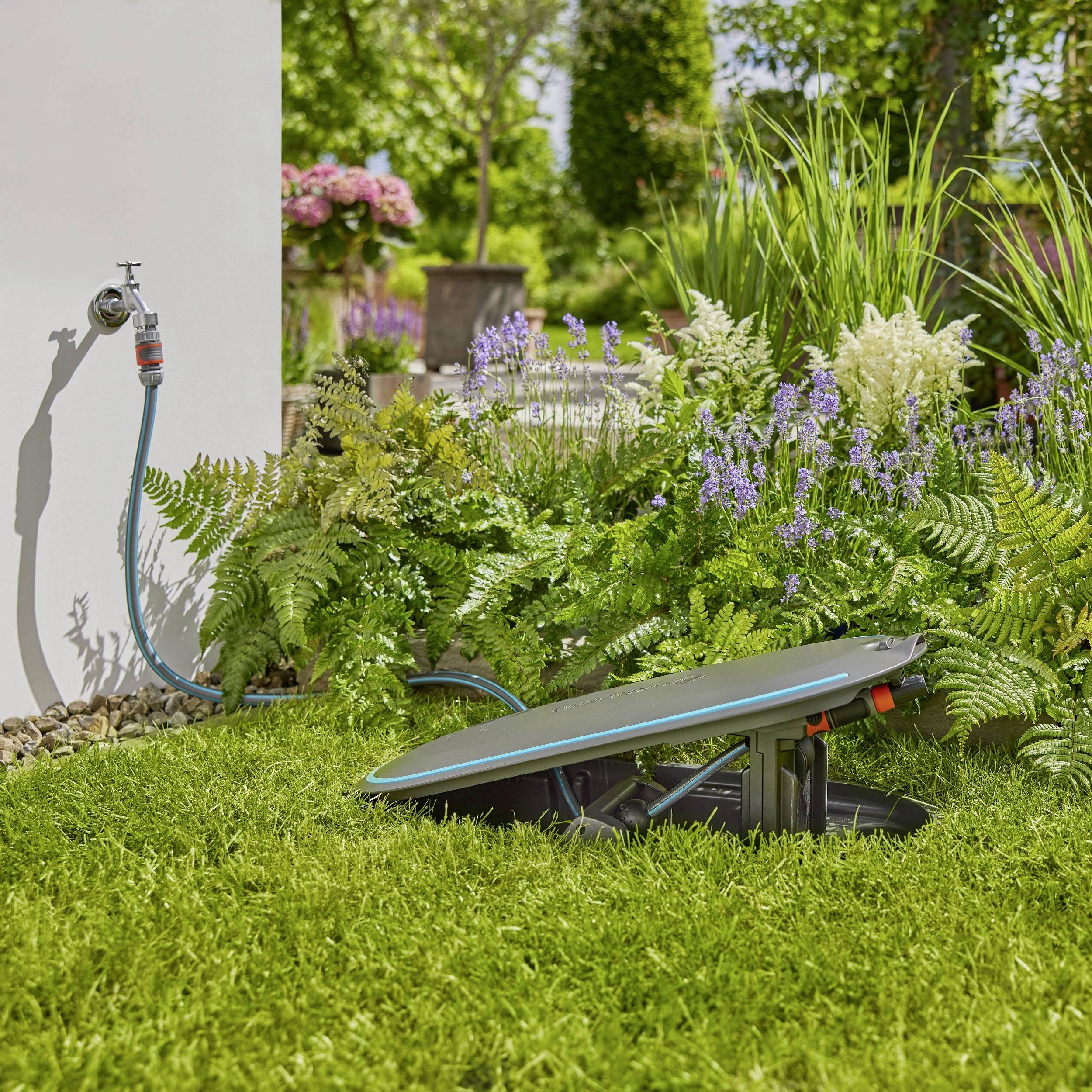 Gazon in de tuin met een verzonken grasmaaier nabij een waterbron, omgeven door planten en bloemen op een zonnige dag.