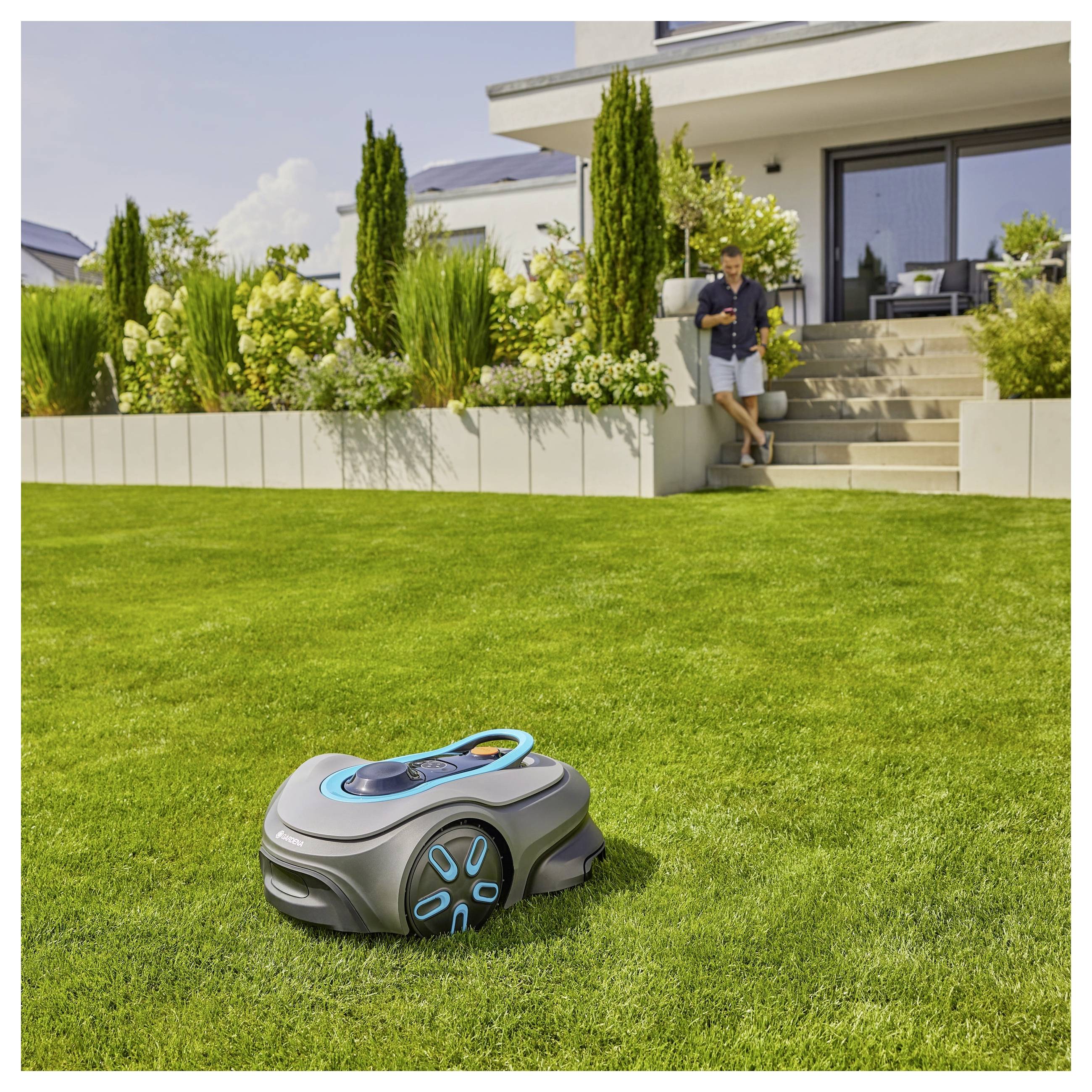 Een robotische grasmaaier snoeit een weelderig groen gazon in een moderne tuin. Op de achtergrond staat een persoon in de buurt van een hedendaags huis.