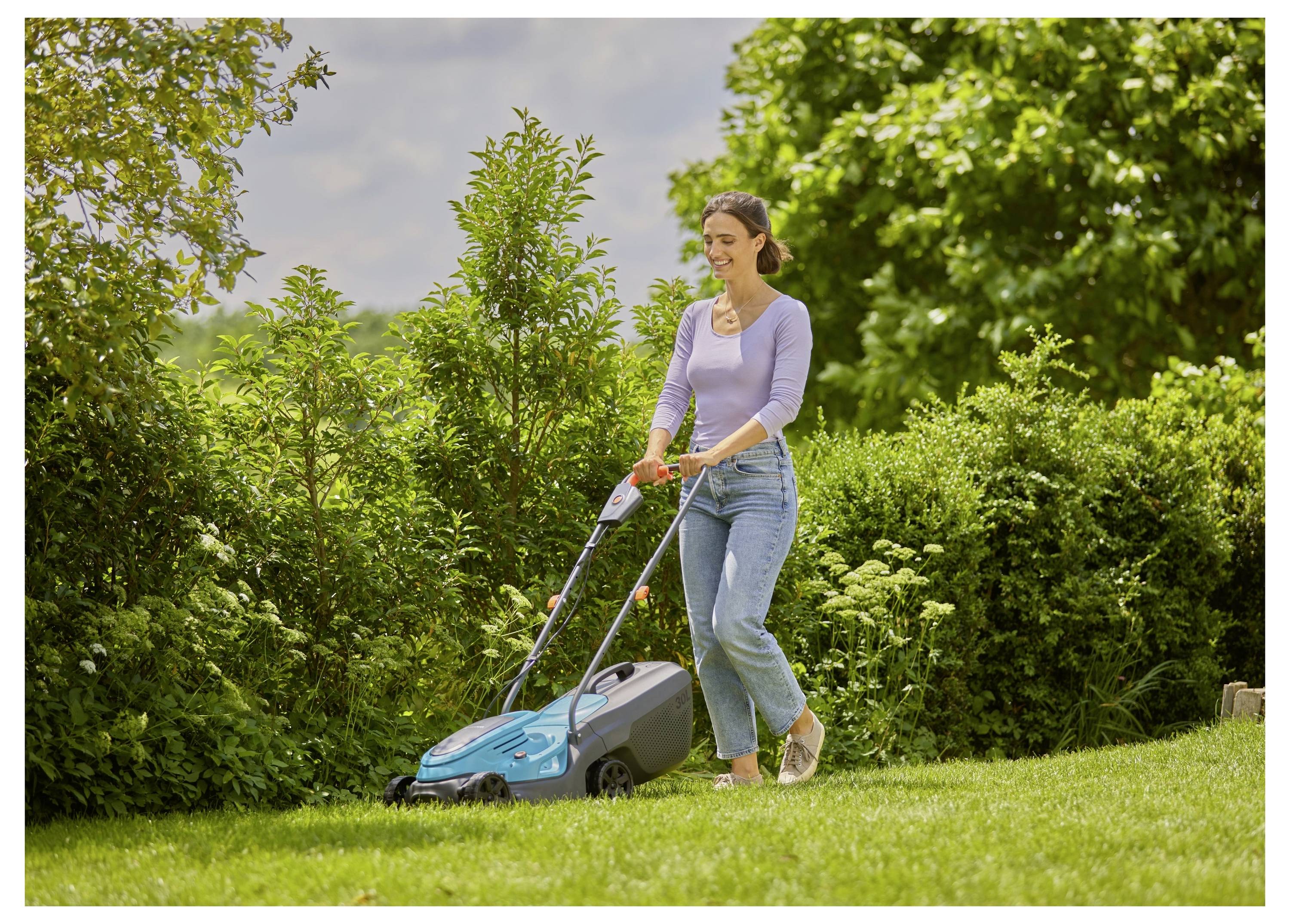 Een vrouw in vrijetijdskleding maait een weelderig groen gazon met een elektrische grasmaaier, omgeven door struiken en bomen op een zonnige dag.