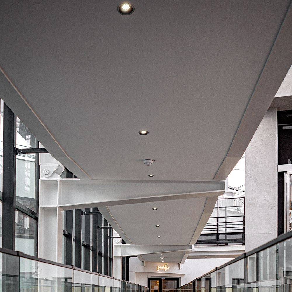 Gang met modern, licht design en glazen balustrade, leidt naar een open ruimte. Bovenaan plafondverlichting, rechts een ingang naar een kamer.