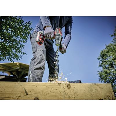 Een persoon boort met een elektrische boor in hout. Houtspaanders vliegen rond. Het is een zonnige dag buiten.