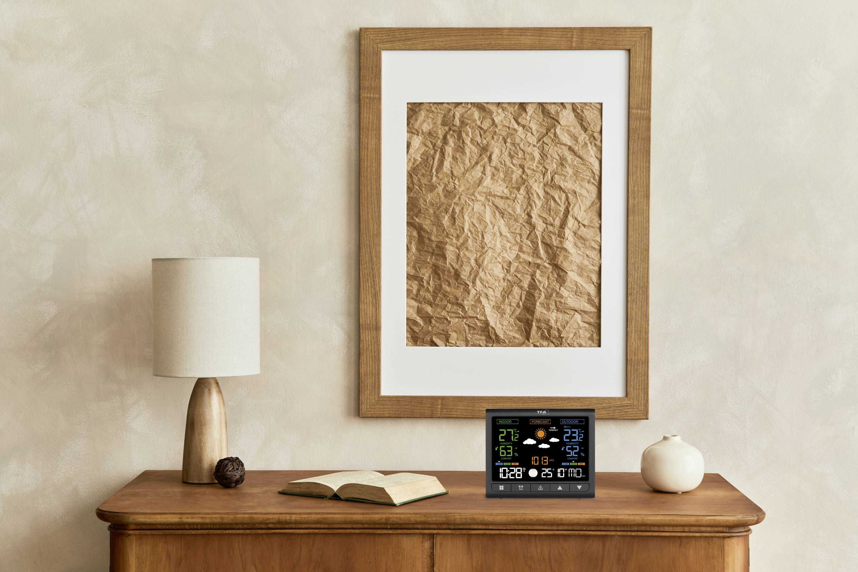 Een houten kast met een lamp, een opengeslagen boek, een kleine pompoenbeeldjes en een digitaal weerstation. Op de achtergrond hangt een kunstwerk aan de muur.