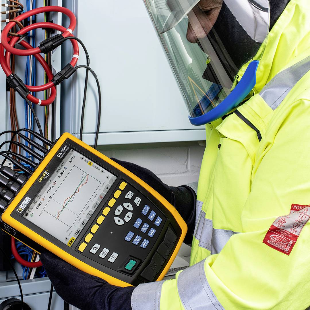 Een technicus in beschermende uitrusting gebruikt een elektronisch meetapparaat aan een bekabelingsinstallatie om gegevens te analyseren.