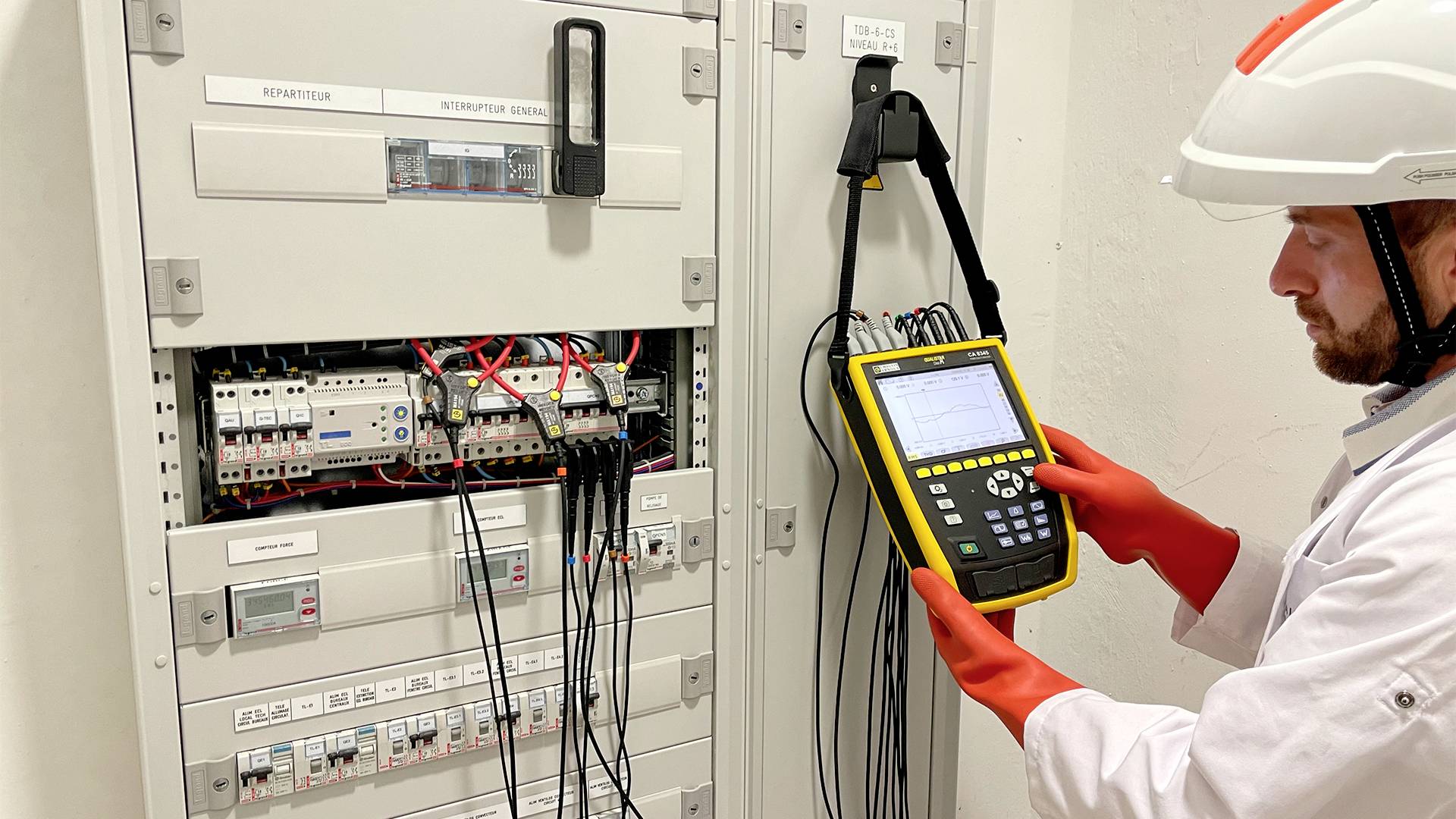 Een technicus in beschermende uitrusting controleert een schakelkast vol kabels en zekeringen met een meetapparaat.