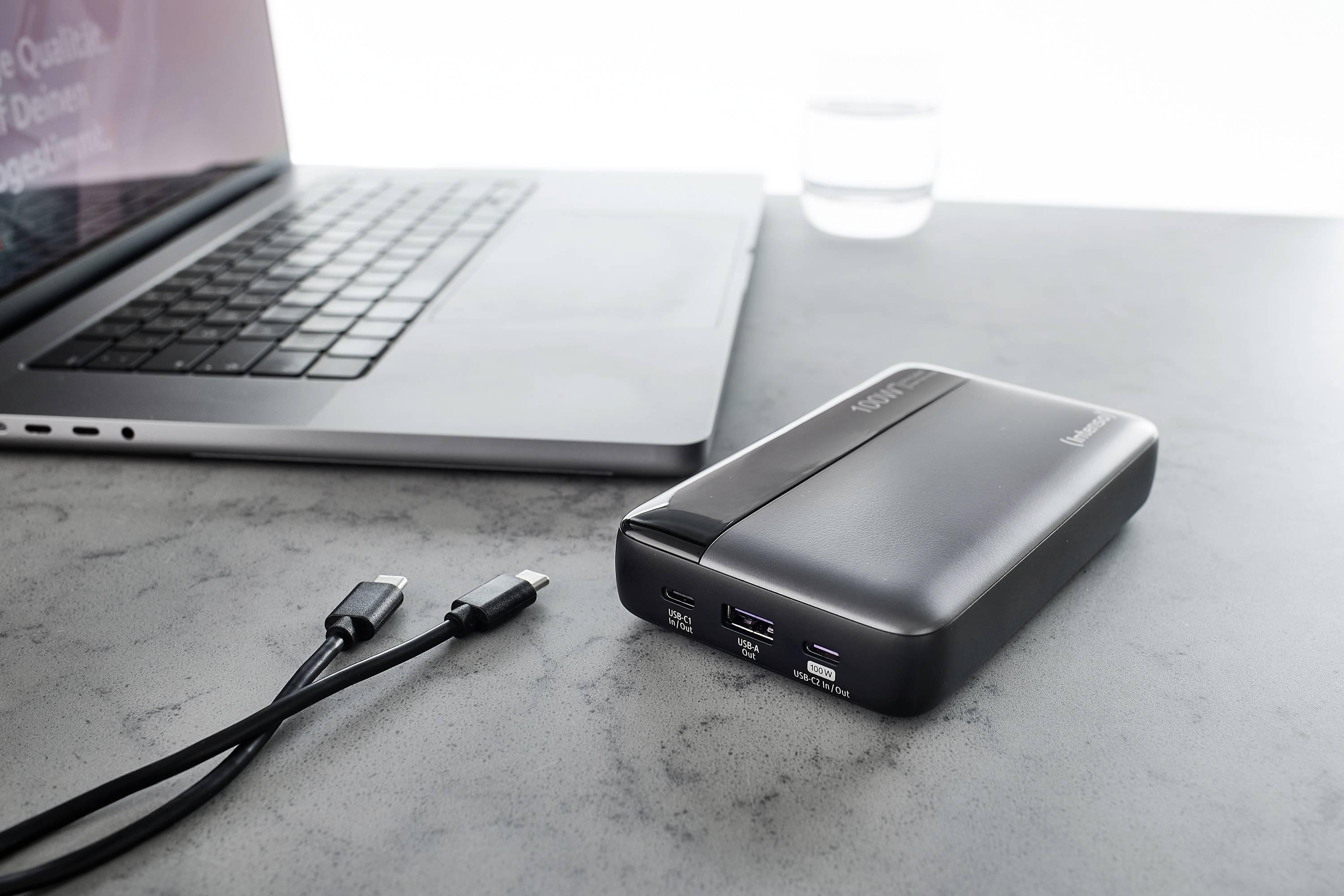 Een powerbank naast een laptop op een grijze tafel, met twee USB-kabels ernaast. Ideaal voor het opladen van apparaten onderweg.