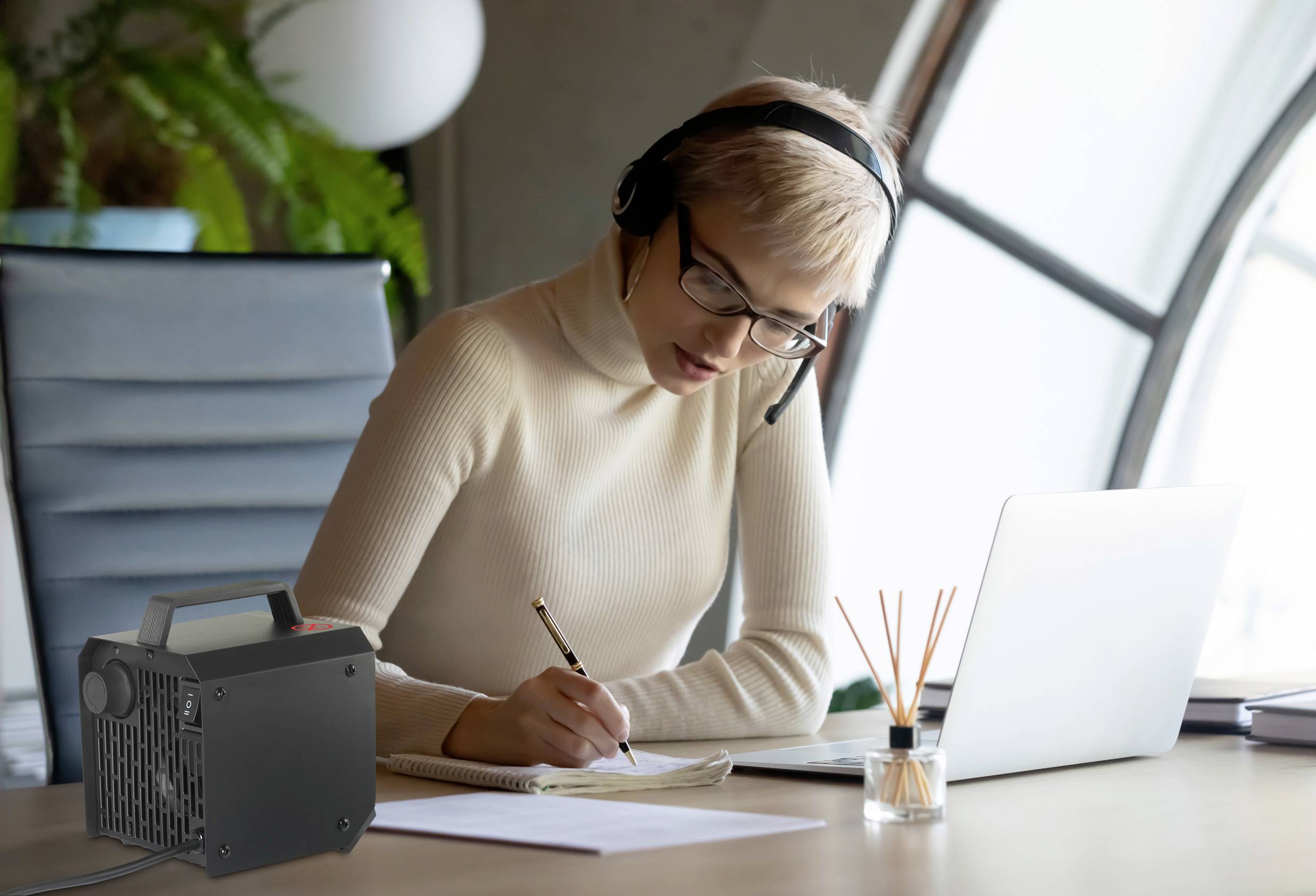 Een persoon met een headset werkt geconcentreerd op een laptop en maakt aantekeningen op papier. Een luchtontvochtiger staat op tafel.
