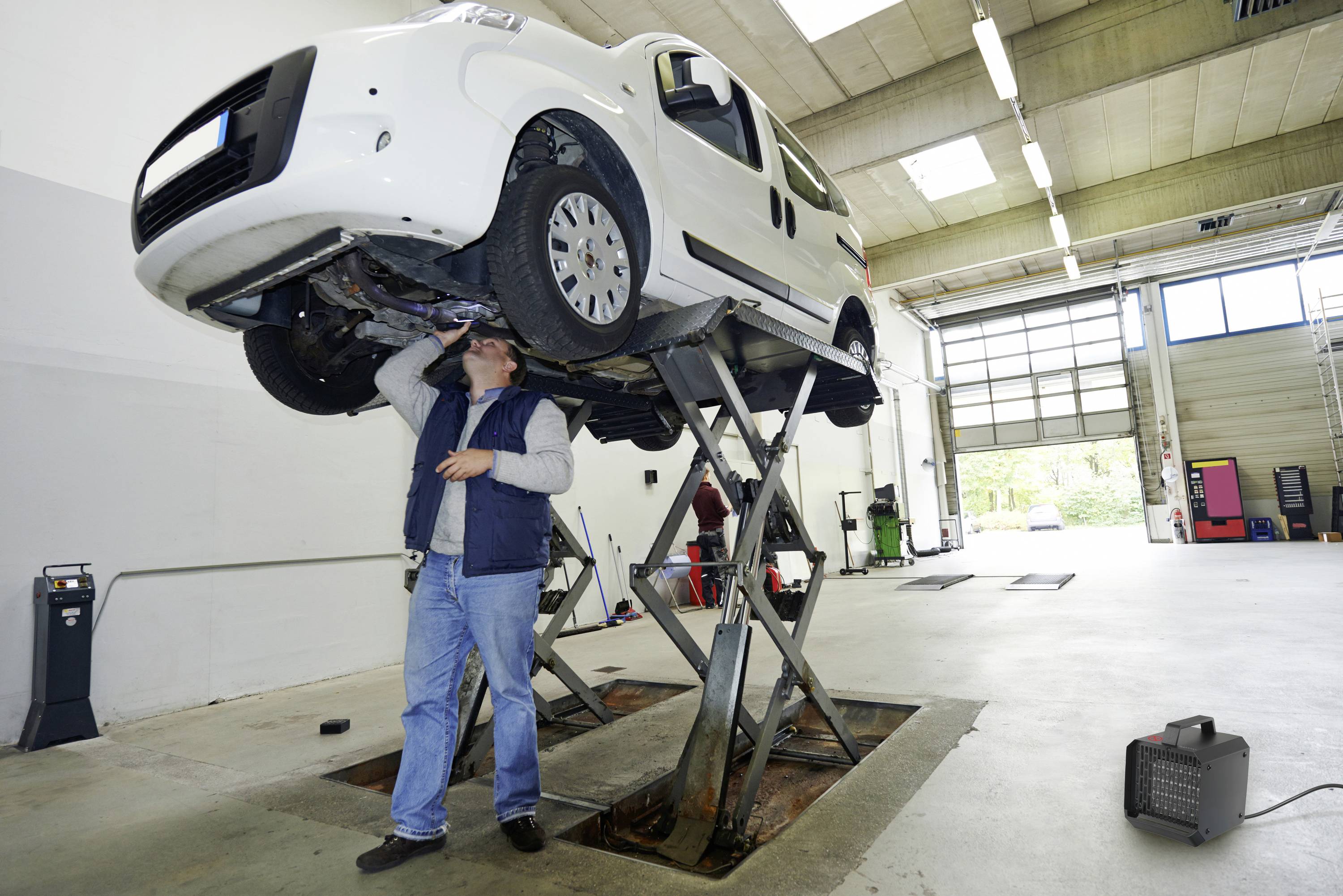 Een monteur inspecteert een witte auto die is opgetild op een hydraulische hefbrug in een garage, waarbij gereedschappen en apparatuur zichtbaar zijn op de achtergrond.