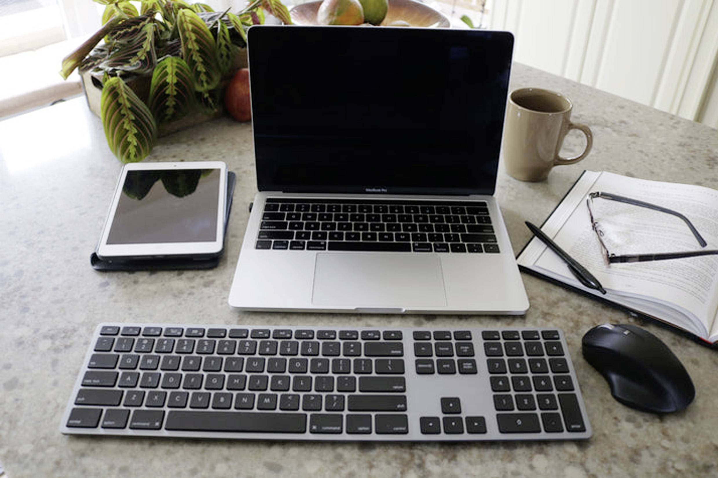 Op een bureau staan een laptop, een tablet, een externe toetsenbord, een muis, een kopje en een opengeslagen notitieboek.