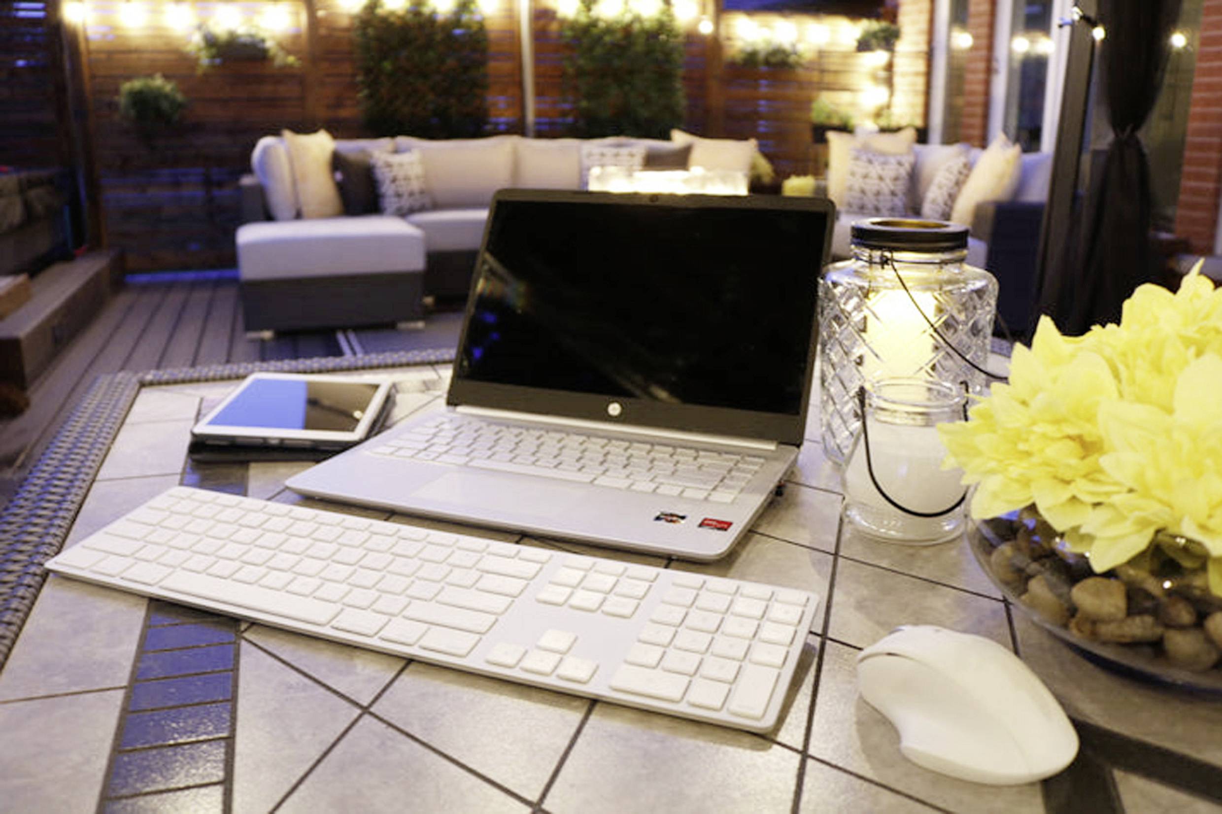 Laptop, tablet, toetsenbord en muis op een terrastafel bij avondverlichting. Omringd door bloemen en een gezellige zithoek.
