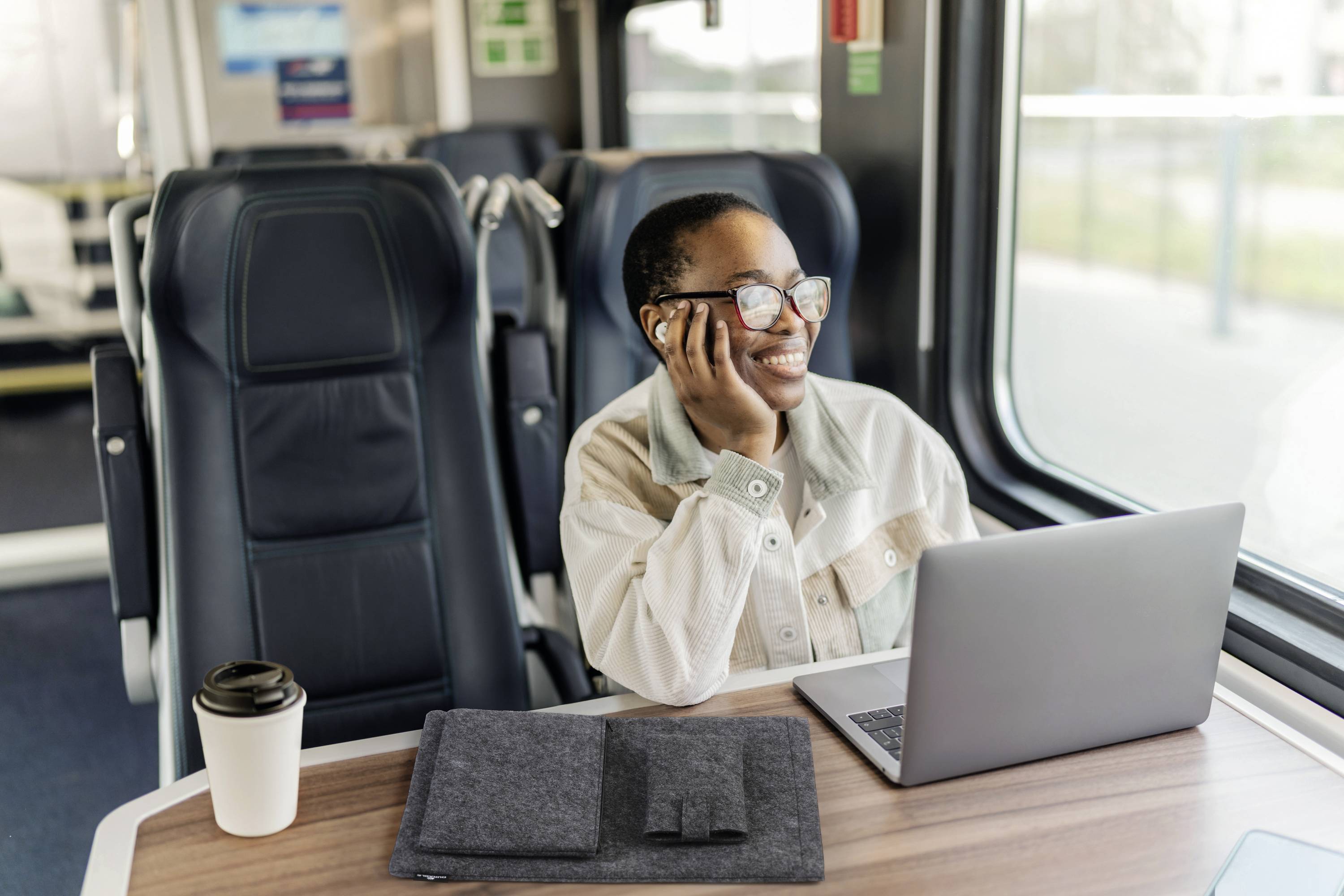 Een persoon zit in een treincoupe, lacht terwijl hij/zij aan het bellen is en kijkt naar een laptop. Een koffiekopje staat op tafel.