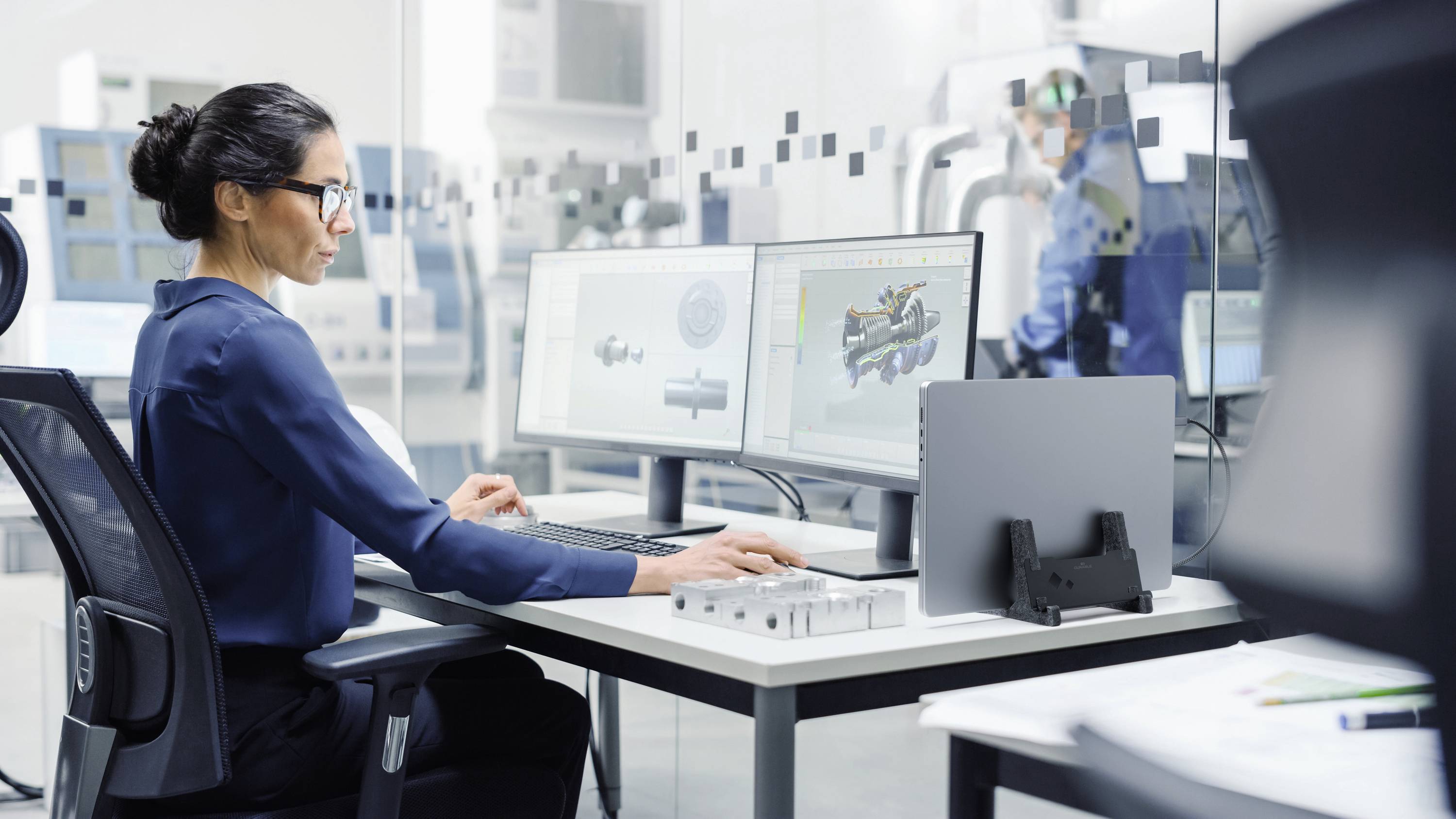 Een vrouw werkt geconcentreerd aan een computer in een moderne industriële omgeving. Op de schermen zijn technische tekeningen zichtbaar.