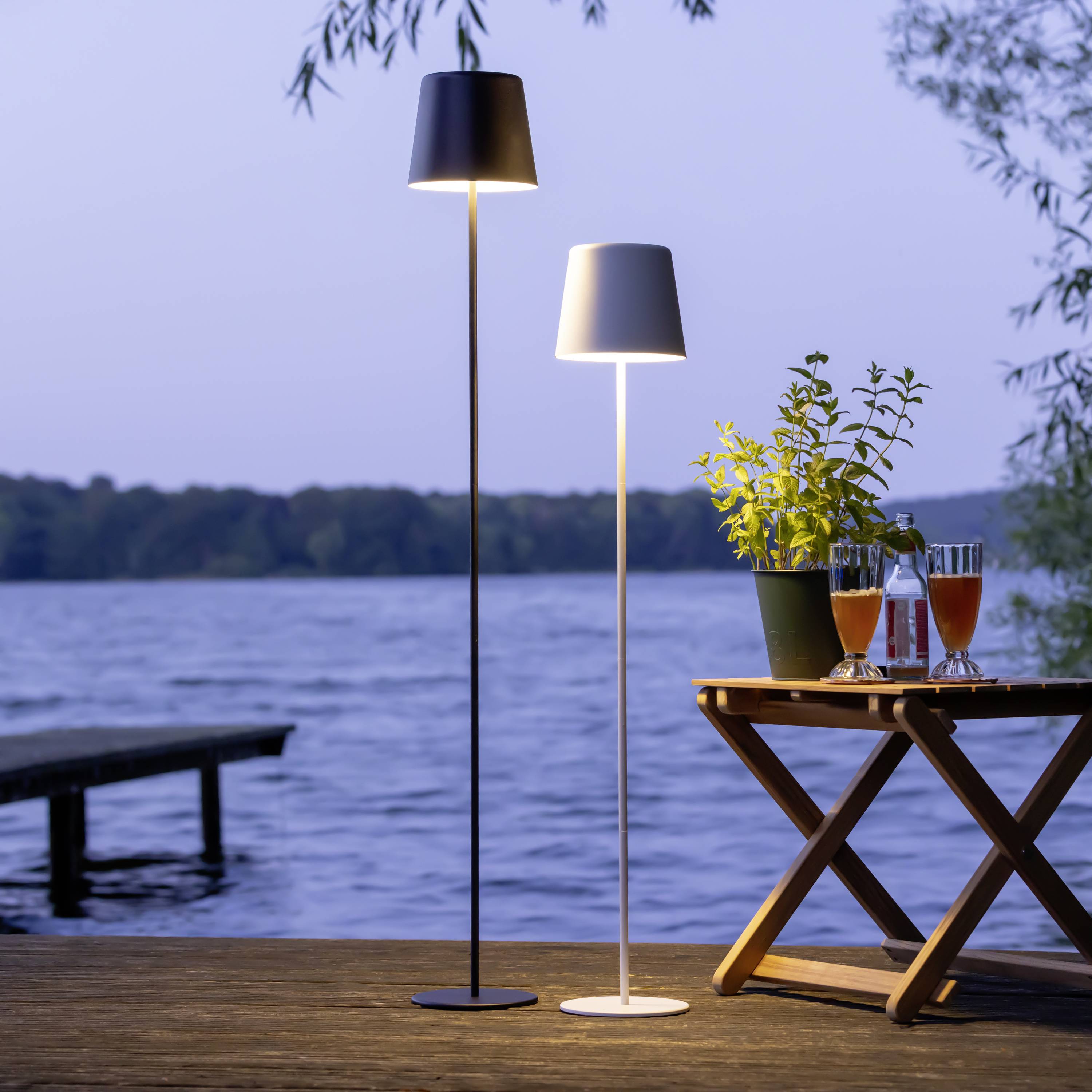 Een tafel met dranken staat op een terras aan het meer, geflankeerd door twee moderne staande lampen. Op de achtergrond is het water zichtbaar.