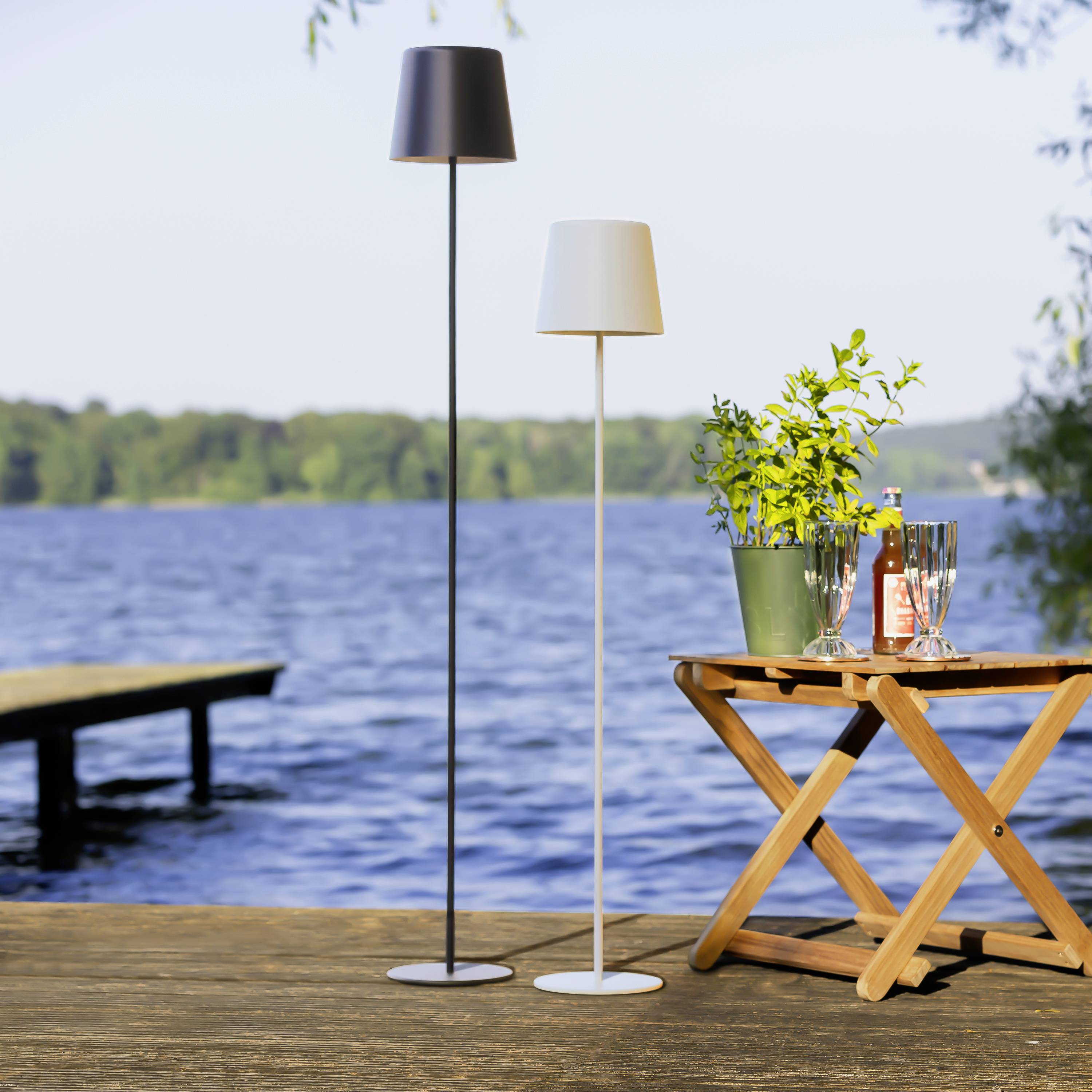Twee staande lampen aan de oever van het meer, een donkere en een lichte, naast een houten tafel met een bloemvaas en dranken. Op de achtergrond een steiger.