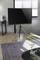 Een modern woonkamer met een televisie op een houten statief. Een donker tapijt bedekt de vloer, naast een salontafel met een plant en een digitale wekker.