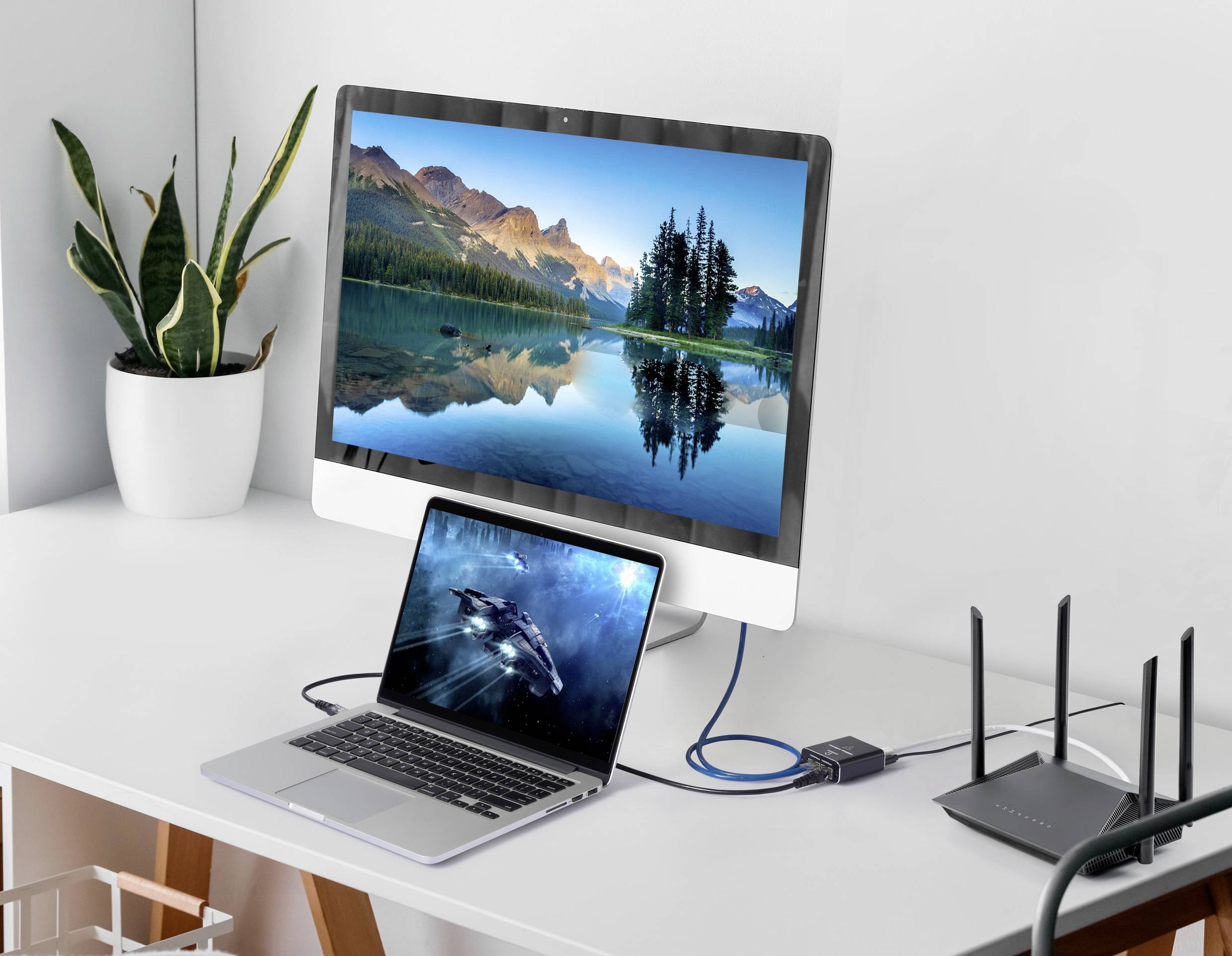 Een bureau met een monitor waarop een landschap te zien is, naast een laptop en een router. De laptop is via een kabel verbonden met de monitor.