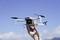 Een hand houdt een kleine drone voor een blauwe lucht. De drone heeft vier propellers en een camera.