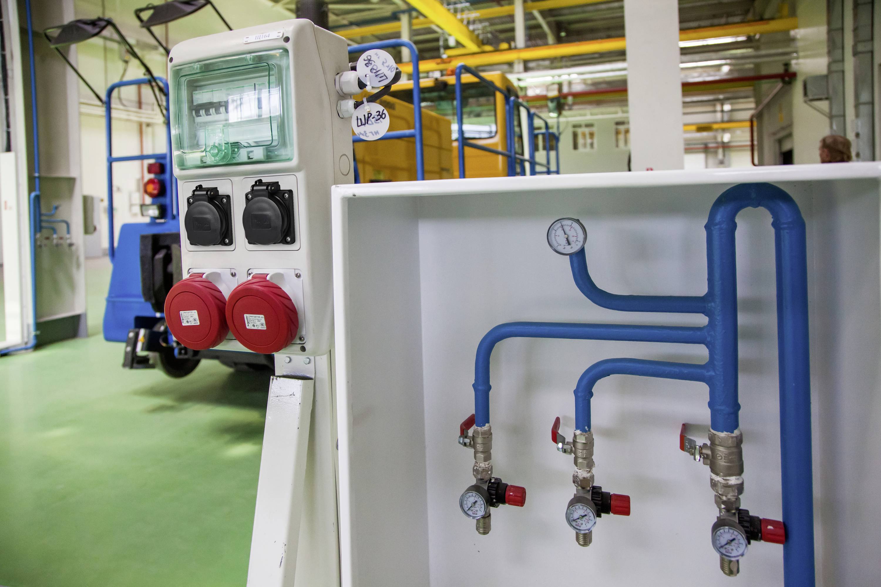 Industriehal met elektrische aansluitingen en blauwe leidingen. Manometers geven druk aan. Gele en blauwe structuur op de achtergrond.