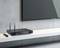 Een zwarte audioreceiver met twee antennes staat op een witte plank onder een aan de muur gemonteerde flatscreen-tv, vergezeld van een afstandsbediening.
