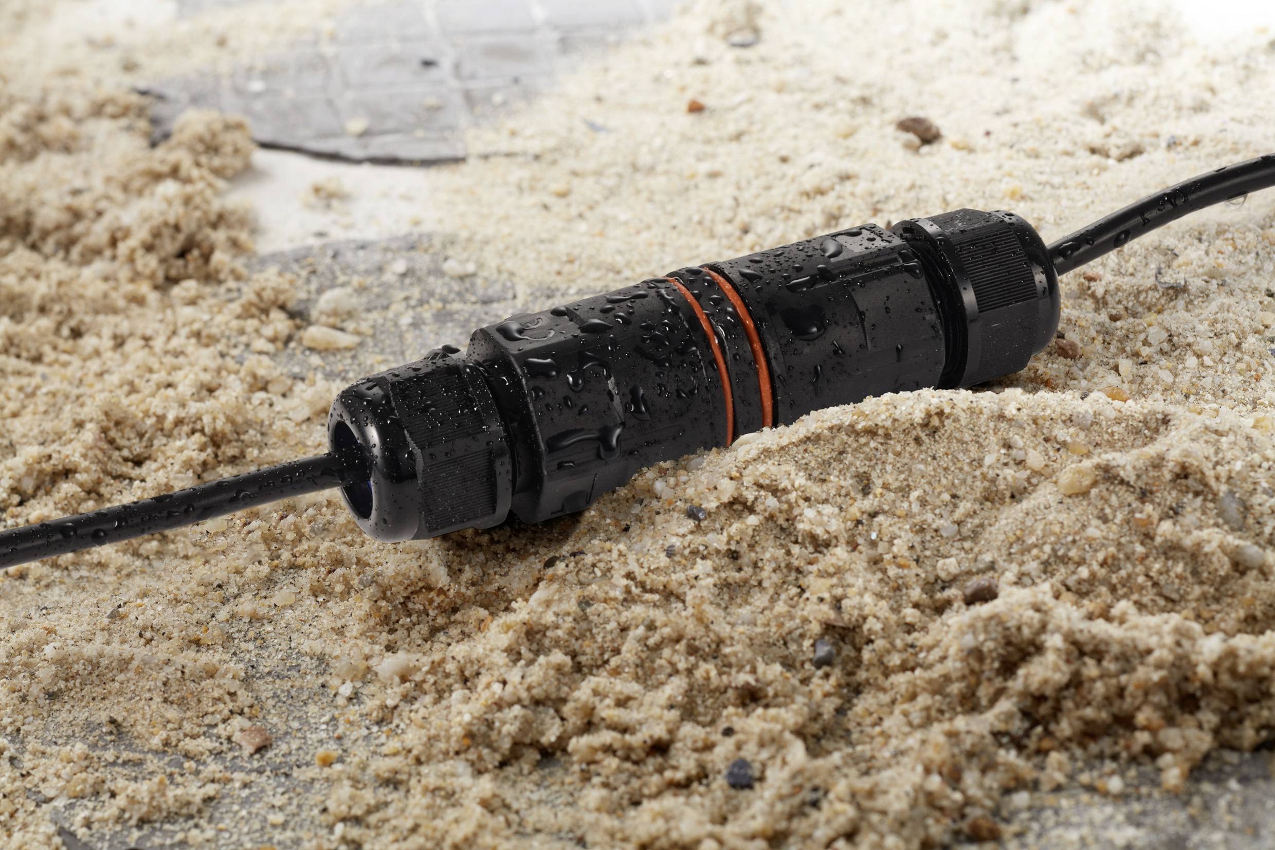 Een waterdichte kabelverbinder rust op een zanderige ondergrond. De kabel is zwart met twee oranje ringen en vertoont waterdruppels.