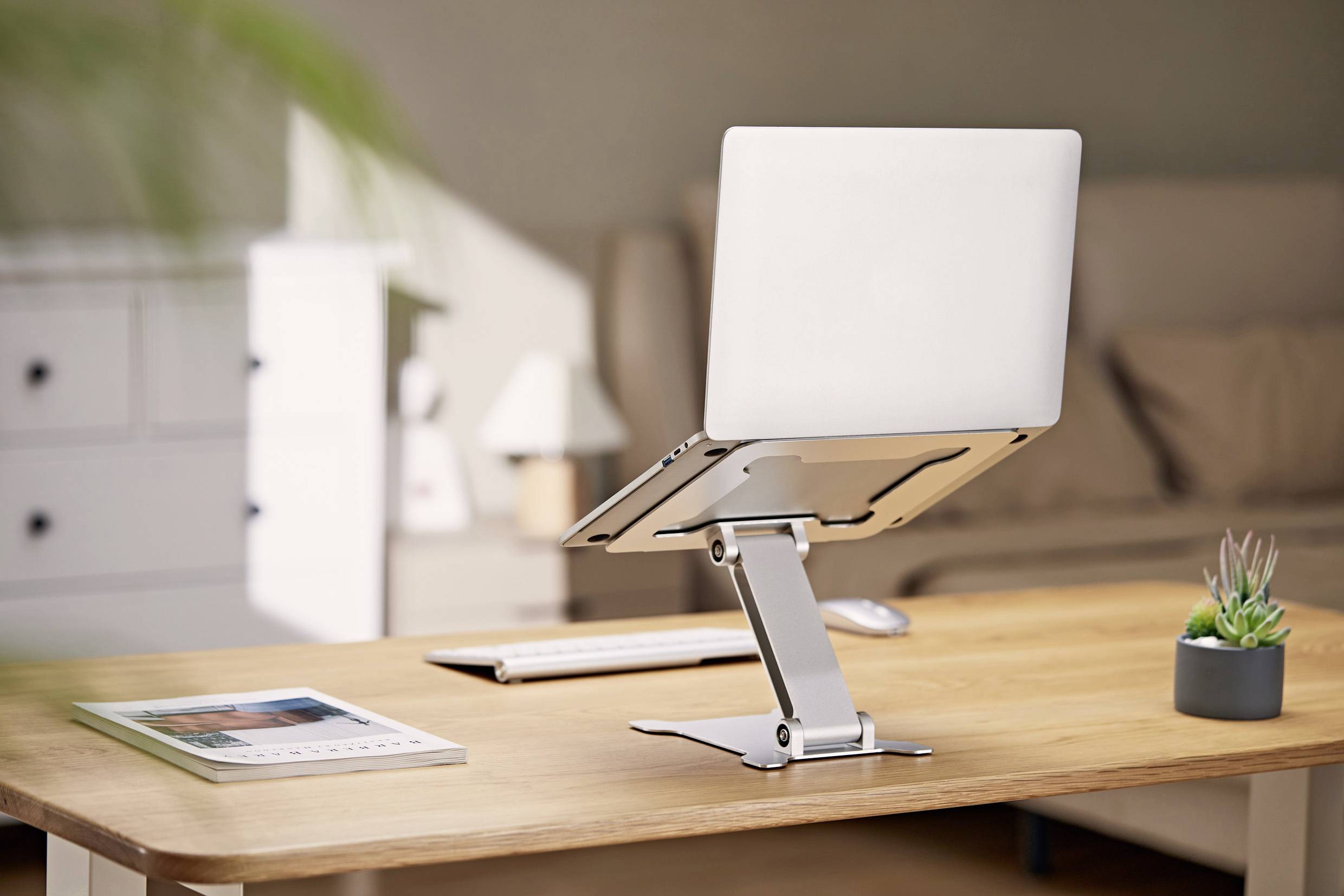 Een laptop staat op een zilveren standaard op een houten tafel. Ernaast liggen een toetsenbord en een muis. Op de achtergrond is een bank.