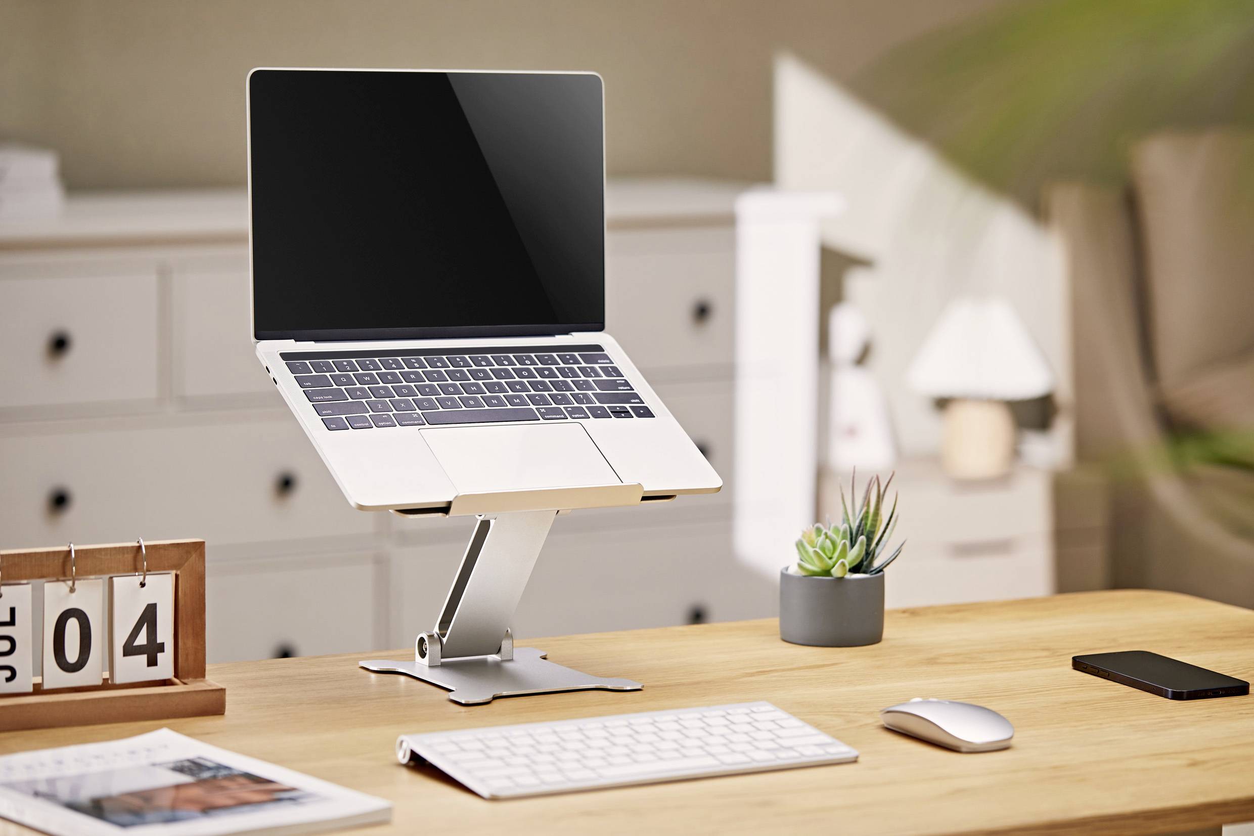 Een moderne bureautafel met een laptop op een standaard, toetsenbord, muis, kalender met datum '04 jul', smartphone en een kleine plant.