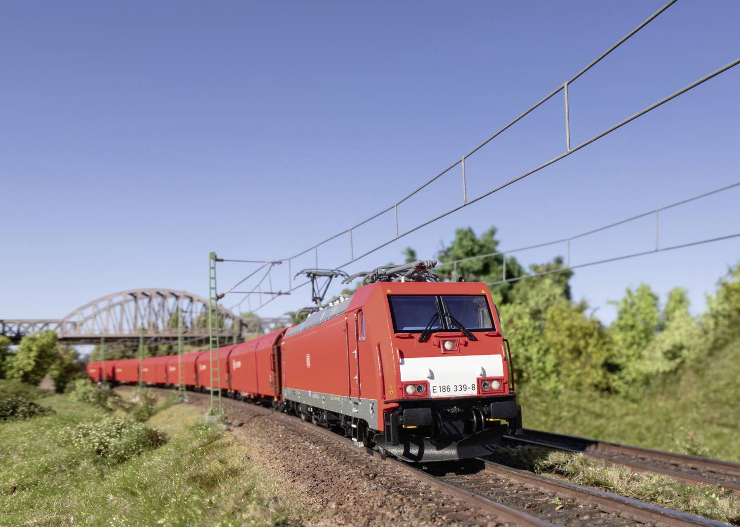 Een rode goederentrein rijdt op een tweesporige route. Op de achtergrond is een brug en enkele bomen te zien.