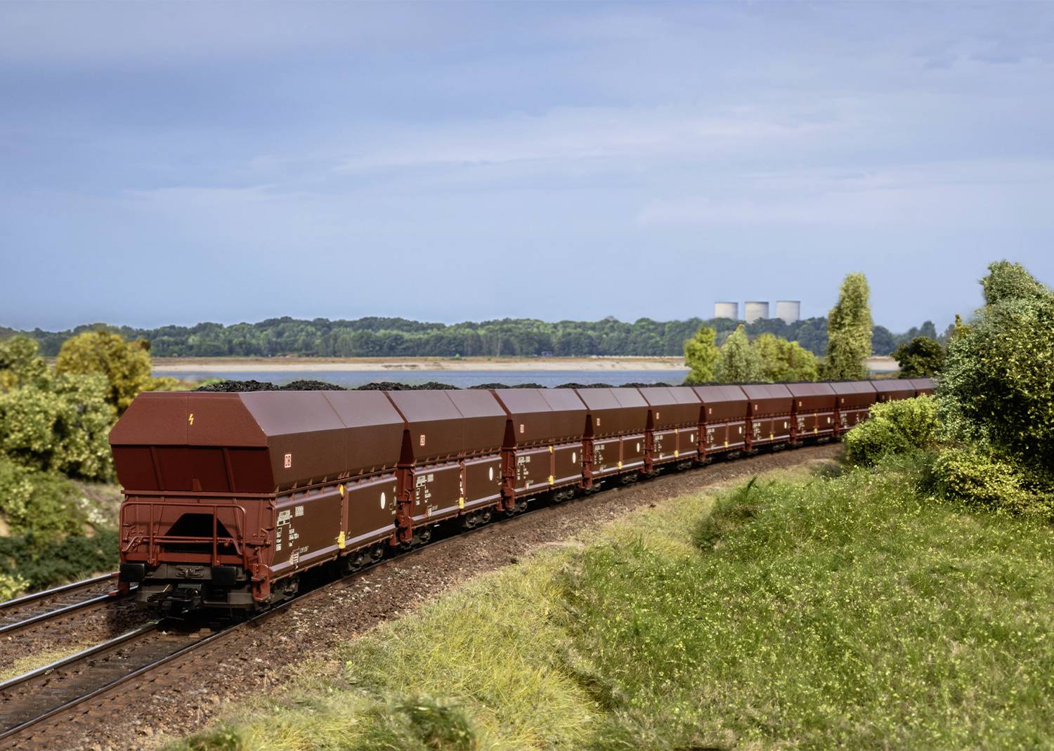 Een lange goederentrein met bruine wagons rijdt over een kronkelige spoorlijn door een groen landschap, met op de achtergrond een elektriciteitscentrale.