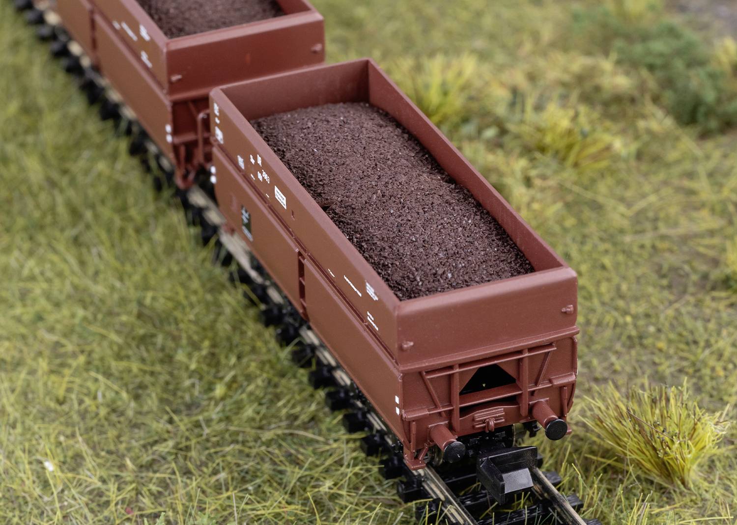 Een modeltrein met bruine lading die rijdt over een spoorweg-achtig miniatuurlandschap met gras. De focus ligt op de trein.