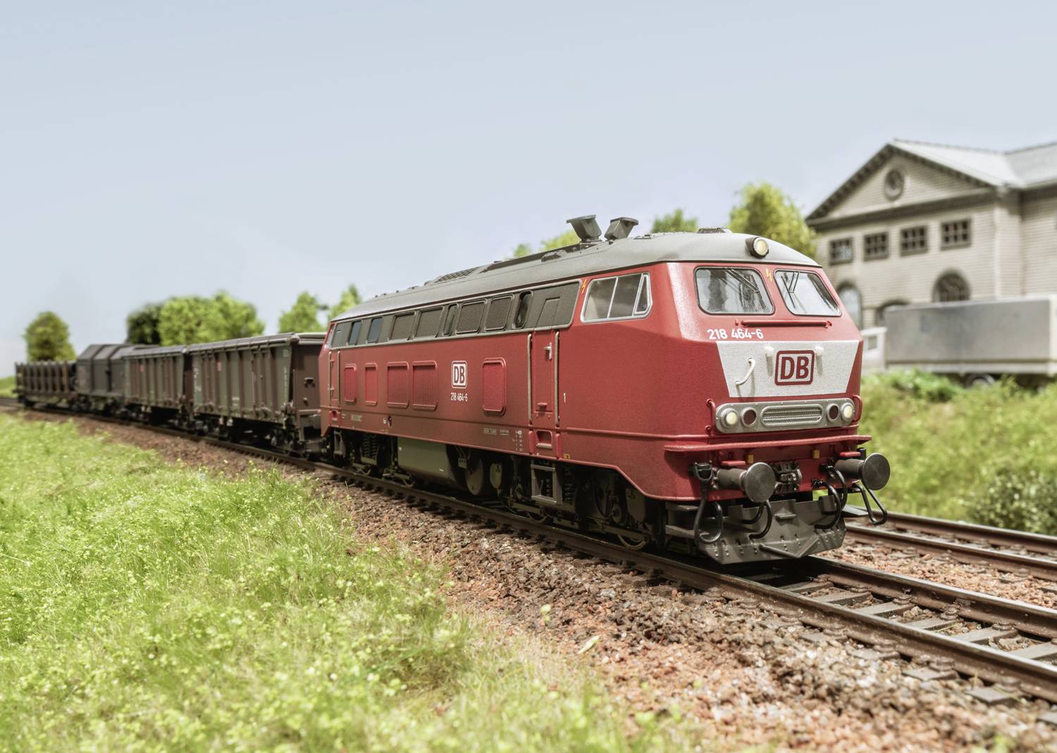 Een rode goederentrein van Deutsche Bahn rijdt over een groene weide in een landelijke omgeving. Op de achtergrond staat een gebouw.