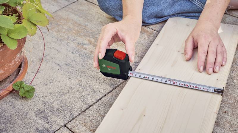 Een persoon meet een houten plank op een terras met een meetlint. Links in het beeld is een plant in een pot te zien.