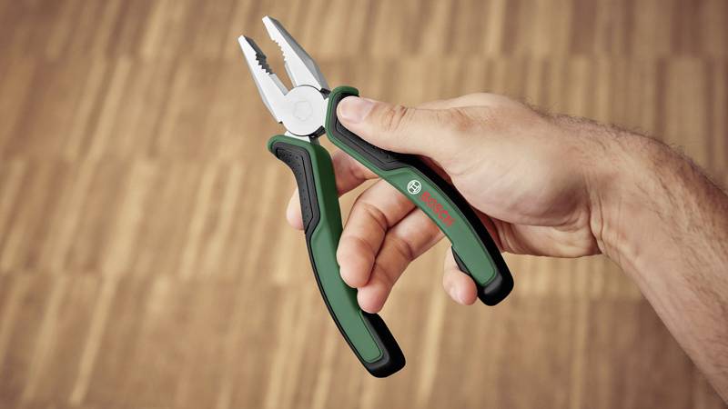Een hand houdt een groen-zwarte tang vast op een lichte houten vloer. De tang draagt het logo van een bekende merknaam.