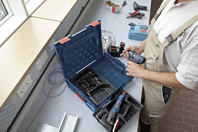Een vakman in werkkleding staat aan een tafel en opent een blauwe gereedschapskoffer met daarin verschillende elektrische gereedschappen.