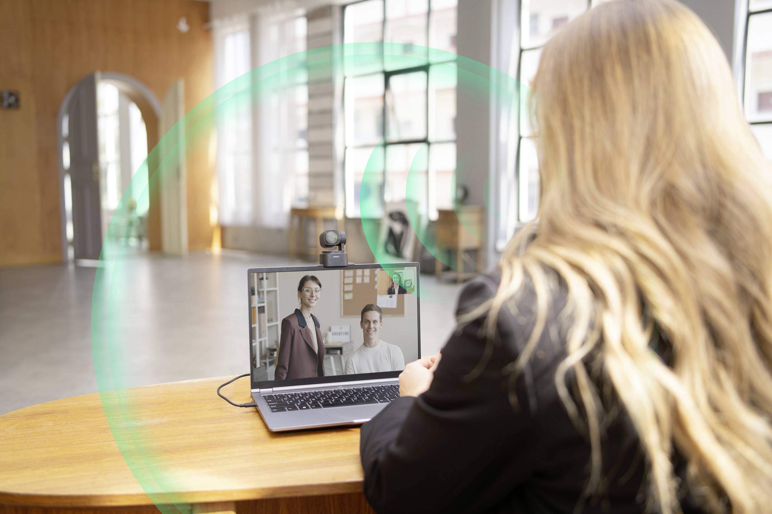 Vrouw voert een videogesprek op een laptop in een kantoor met grote ramen. Twee personen zijn zichtbaar op het scherm.