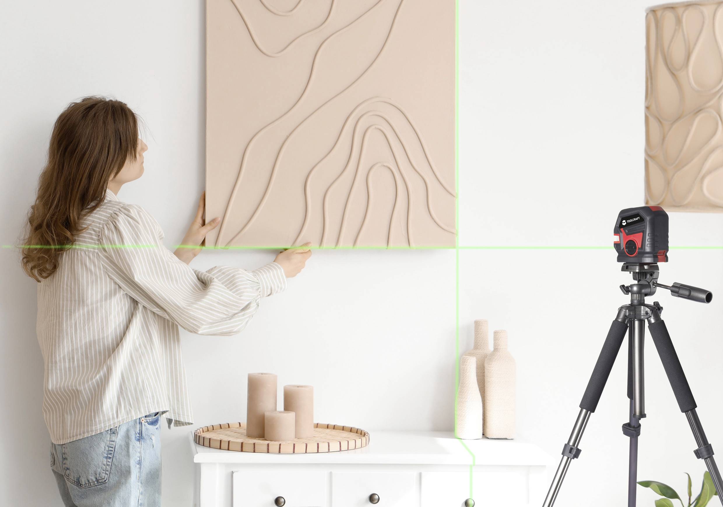 Een vrouw hangt een kunstwerk met golvend patroon aan de muur, terwijl een laserniveaumeter op een statief de uitlijning controleert.