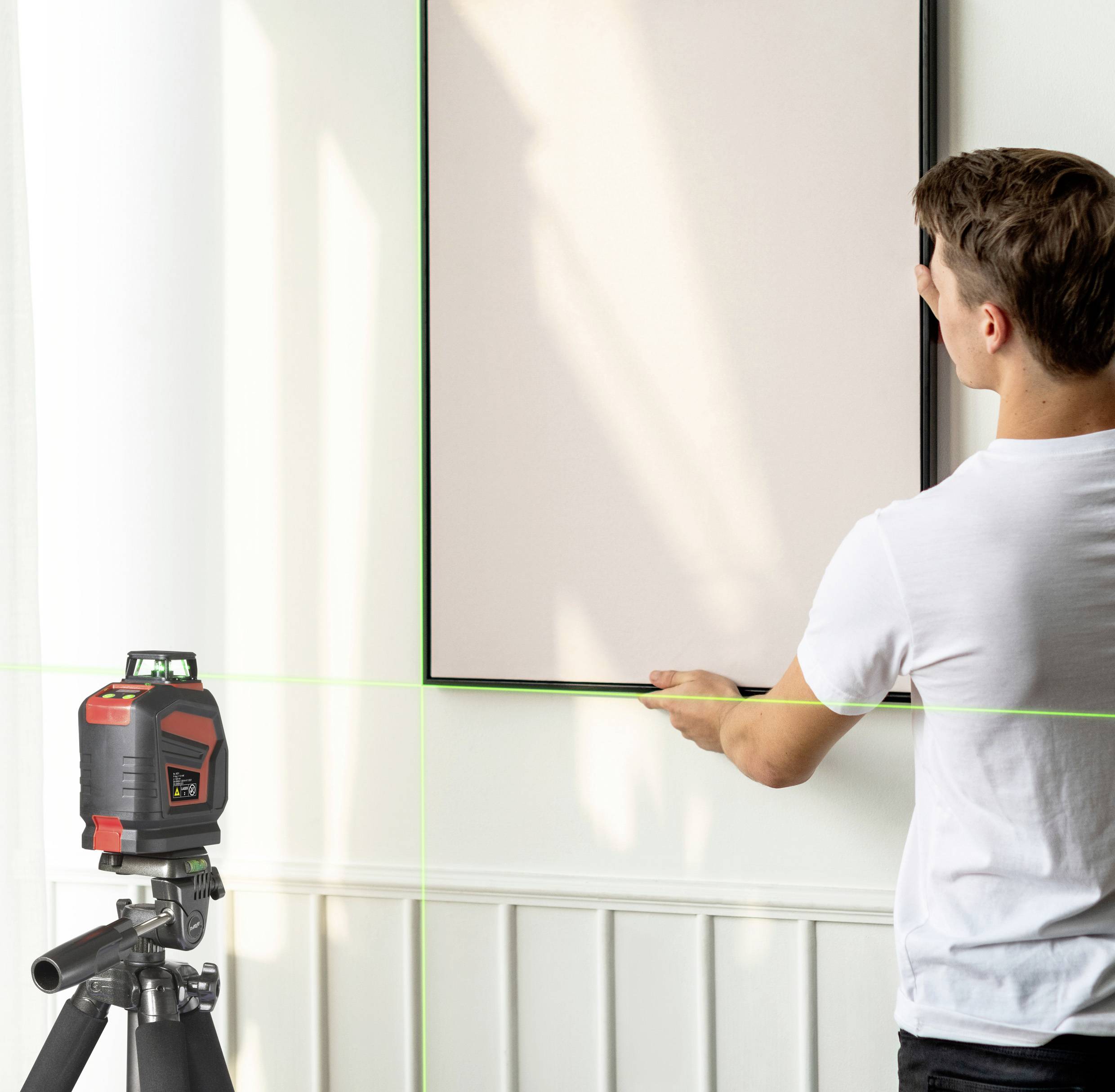 Een man richt een lasertoestel op een muur in een ruimte. Het apparaat projecteert groene lijnen die worden gebruikt voor uitlijning.