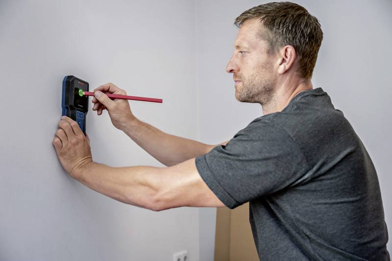 Een man gebruikt een elektronisch meetapparaat en een rode pen op een witte muur, waarschijnlijk voor het uitlijnen of boren.