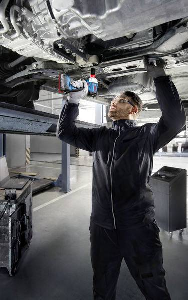 Een persoon in werkkleding repareert met een gereedschap een auto van onderen in een werkplaatsomgeving.