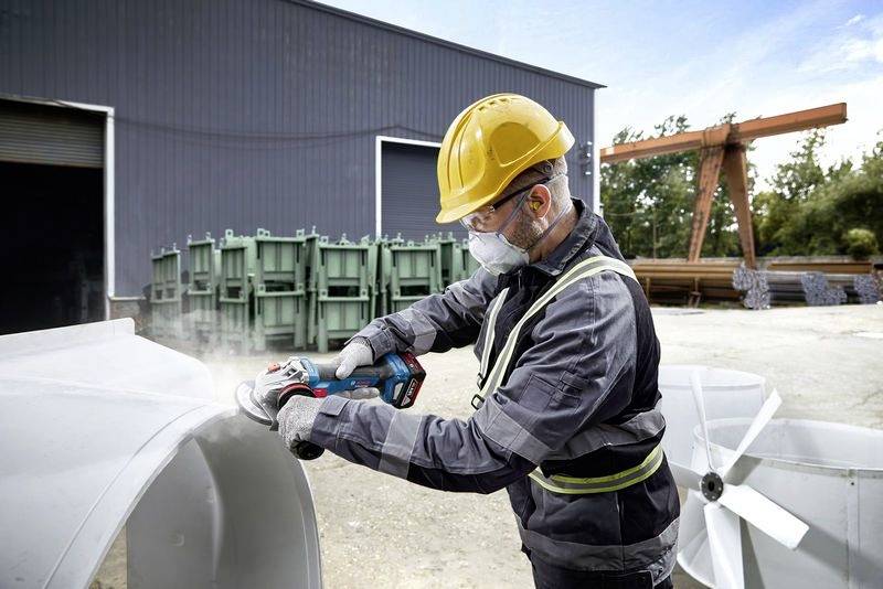 Een arbeider in beschermende uitrusting gebruikt een elektrisch gereedschap om metaal te snijden in een industrieel gebied met machines op de achtergrond.