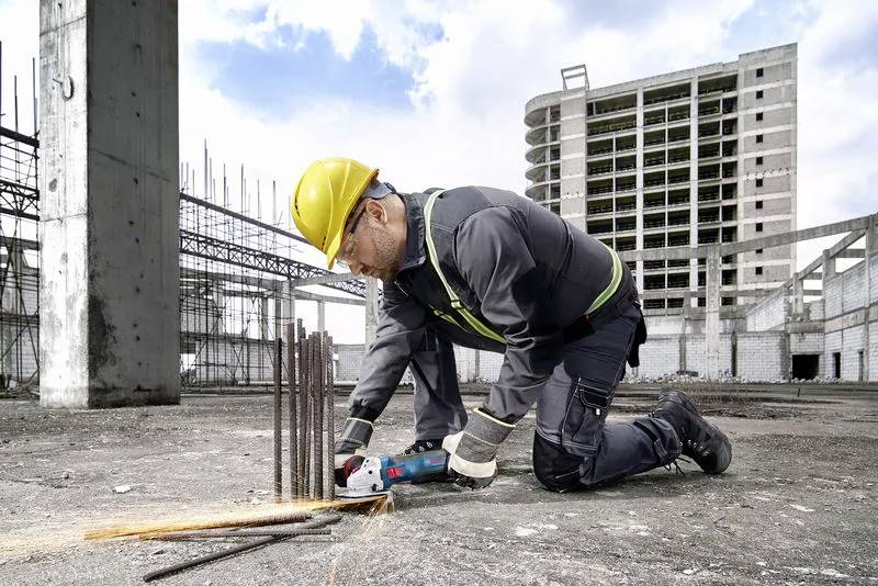 Een bouwvakker in veiligheidsuitrusting snijdt metalen staven door met een elektrisch gereedschap op een bouwterrein, met op de achtergrond een gedeeltelijk opgetrokken hoogbouw.
