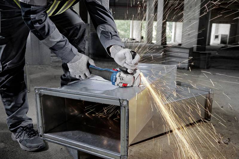 Een persoon snijdt een metalen kanaal met een haakse slijper, waarbij vonken rondvliegen. De achtergrond van een bouwplaats duidt op renovatiewerkzaamheden.