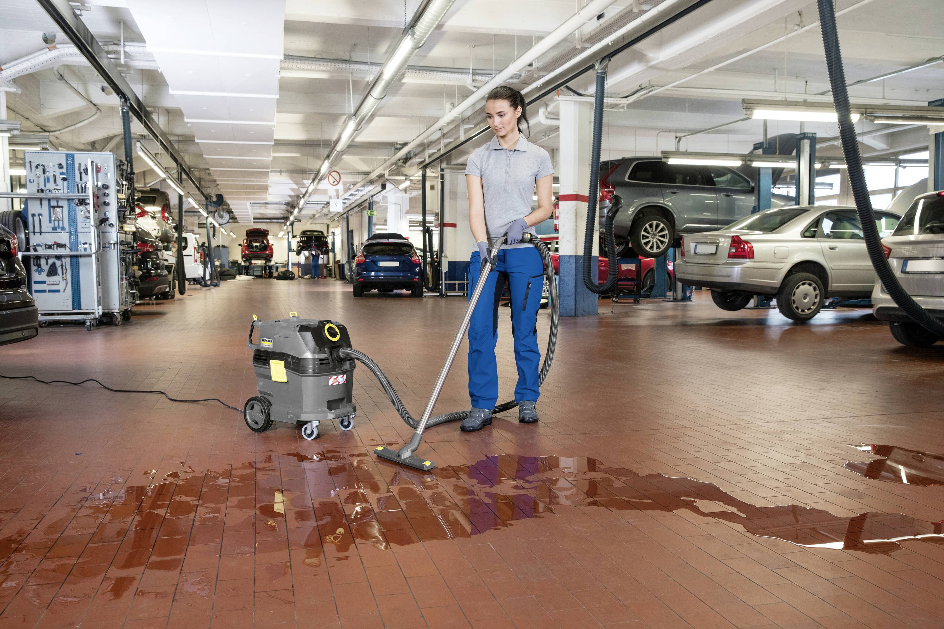 Een persoon reinigt de vloer in een autogarage met een nat-stofzuiger om waterplassen te verwijderen. Op de achtergrond zijn auto's zichtbaar.