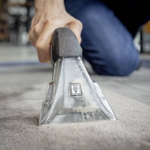 Een persoon reinigt een tapijtoppervlak met een handtapijtcleaner, gefocust op de reinigingsnozzle en het opzuigen van vuil.