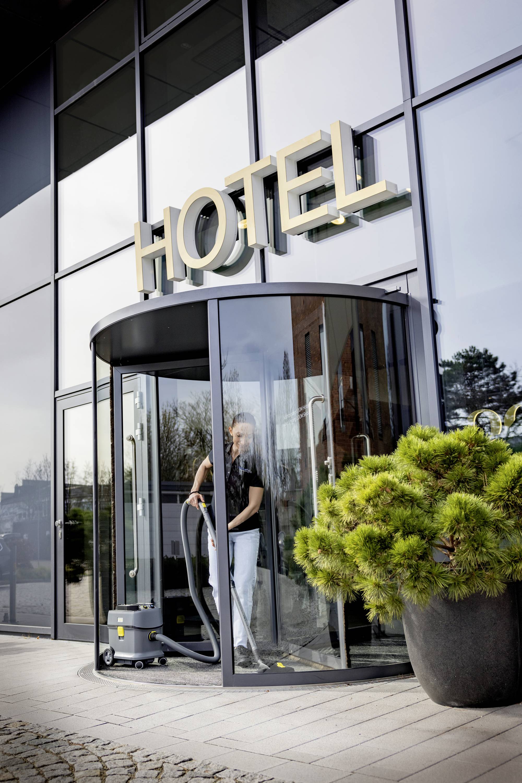 Een persoon reinigt de ingang van een modern hotel met glazen deuren. Grote gouden letters boven de deur vormen het woord 'HOTEL'.