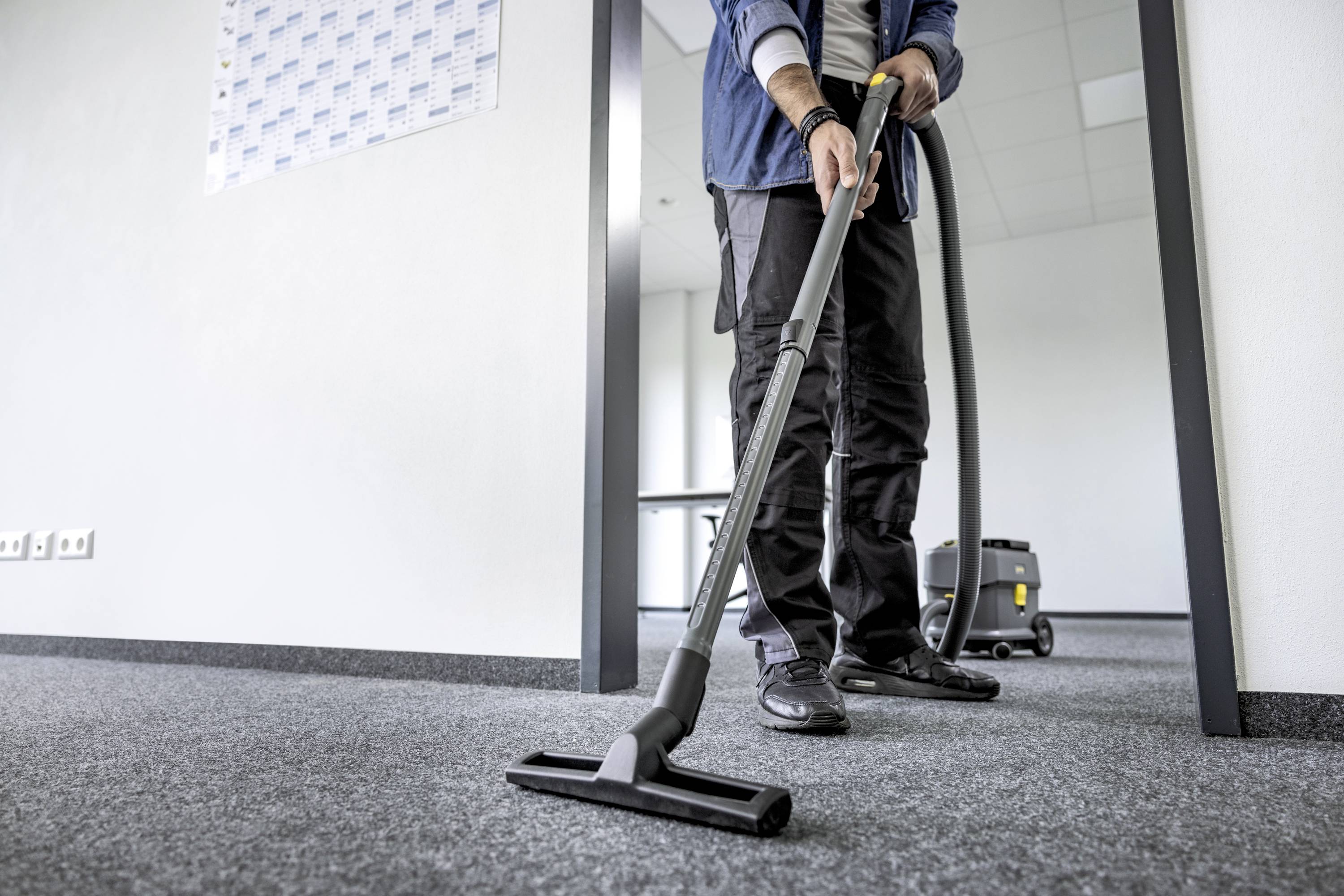 Persoon die een stofzuiger gebruikt om de tapijtvloer in een kantoor schoon te maken. Op de achtergrond zijn meubels en een muur zichtbaar.