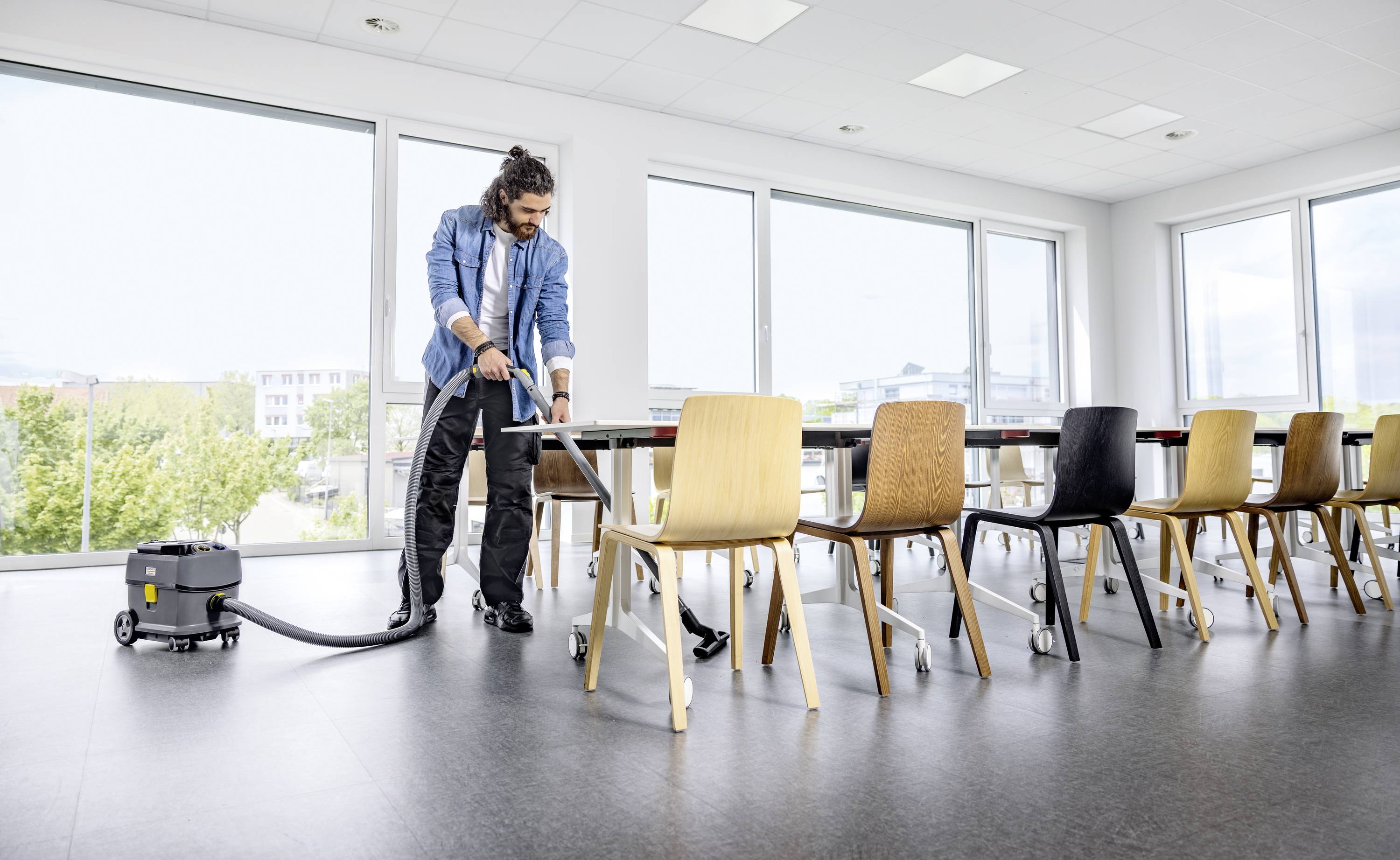 Een persoon reinigt de vloer van een vergaderzaal met een stofzuiger. Lichte stoelen staan rond een lange tafel met grote ramen op de achtergrond.